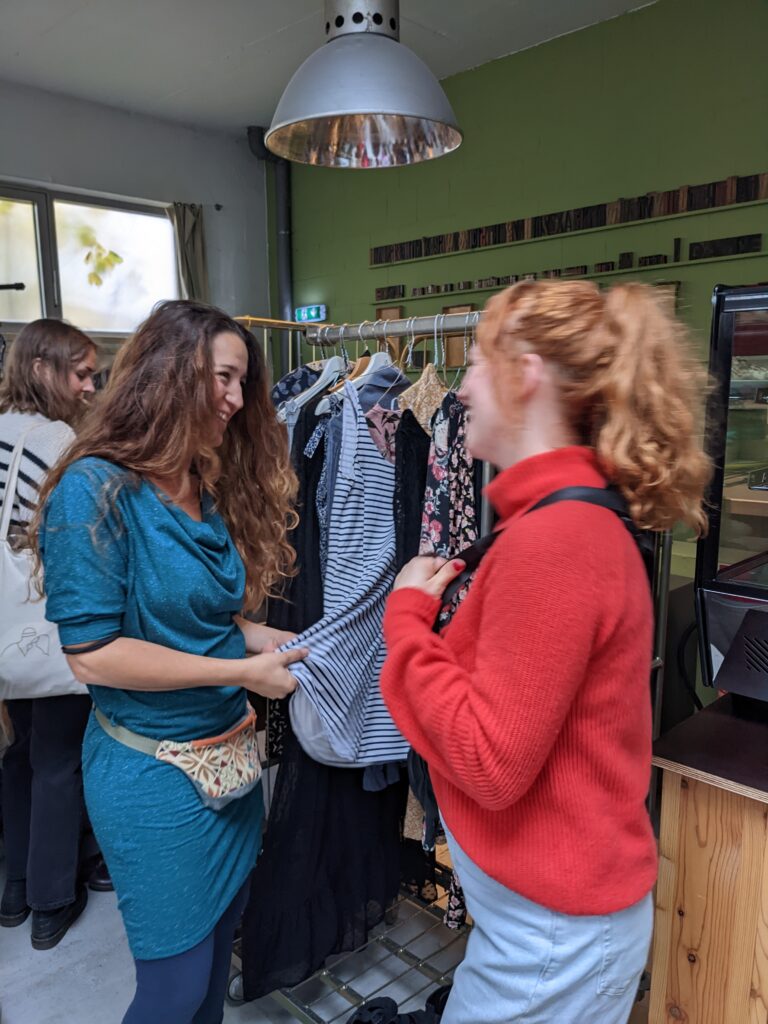 Zwei Frauen stehen vor einem Kleiderständer mit Kleidern und lachen