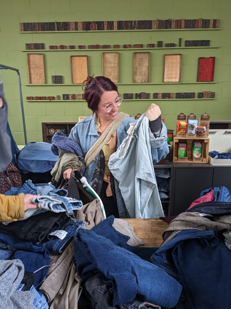 Frau steht an einem Tisch, der vollgepackt ist mit Hosen, fast ausschließlich Jeans. Sie trägt Kleidung über dem Arm und geht durch die Hosen