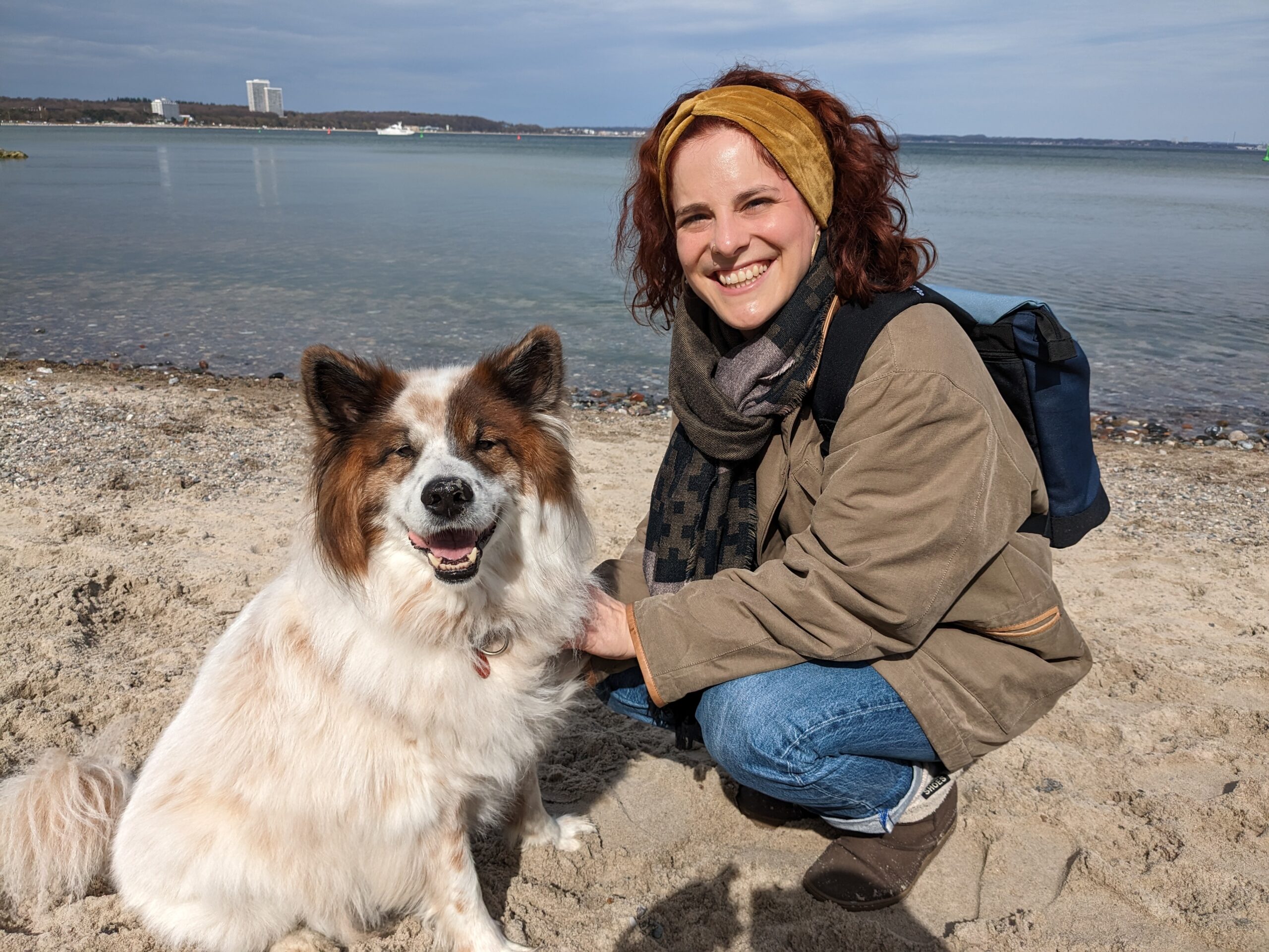 Frau mit roten Locken lächelt am Wasser mit ihrem Hund